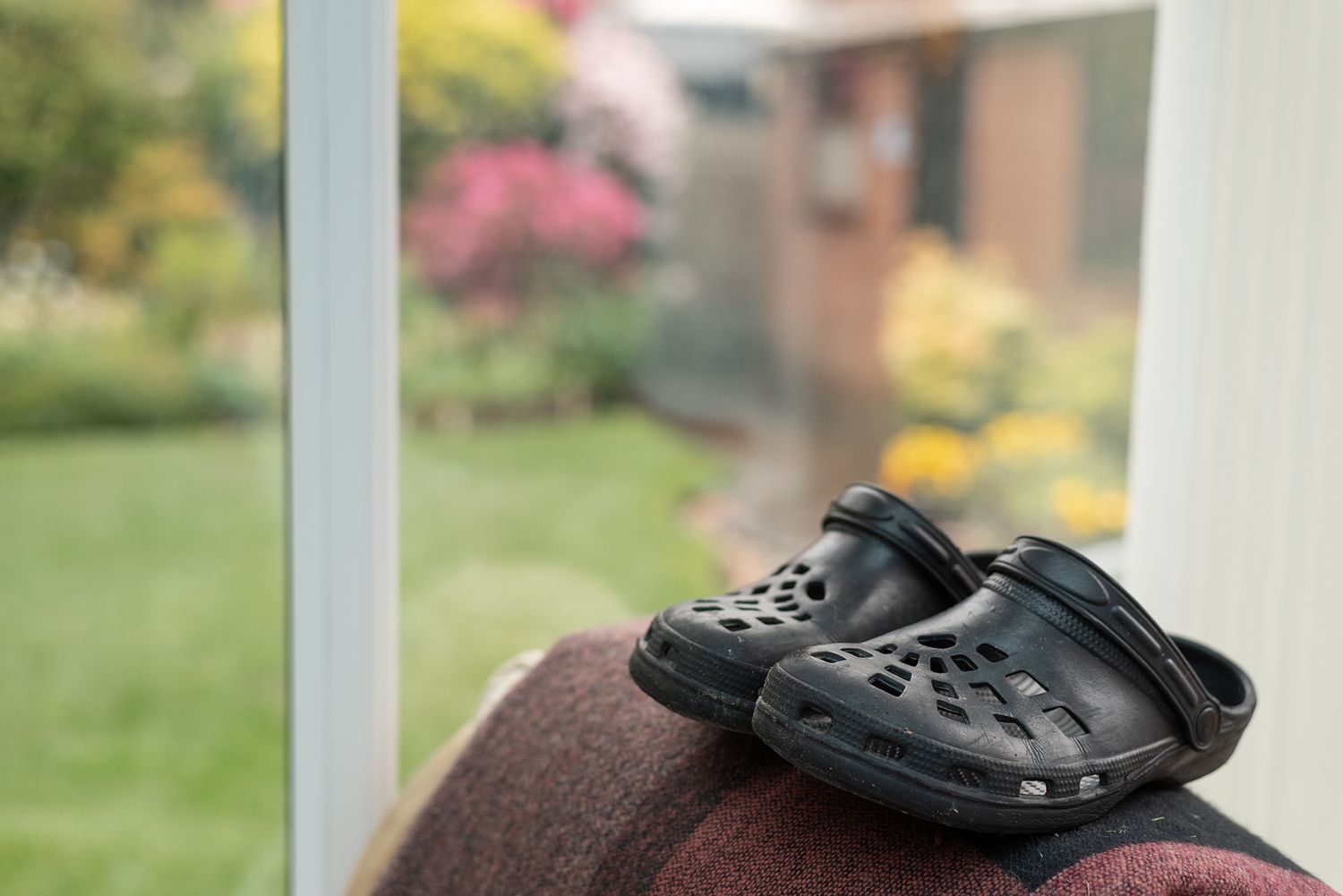 Black Crocs clogs resting on a chair back near a window with a colorful garden view.