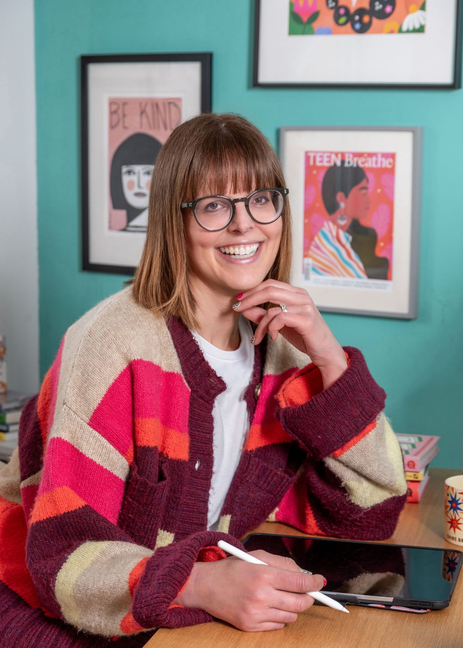 Smiling woman in striped cardigan at desk with colorful art prints and Teen Breathe magazine poster on teal wall.