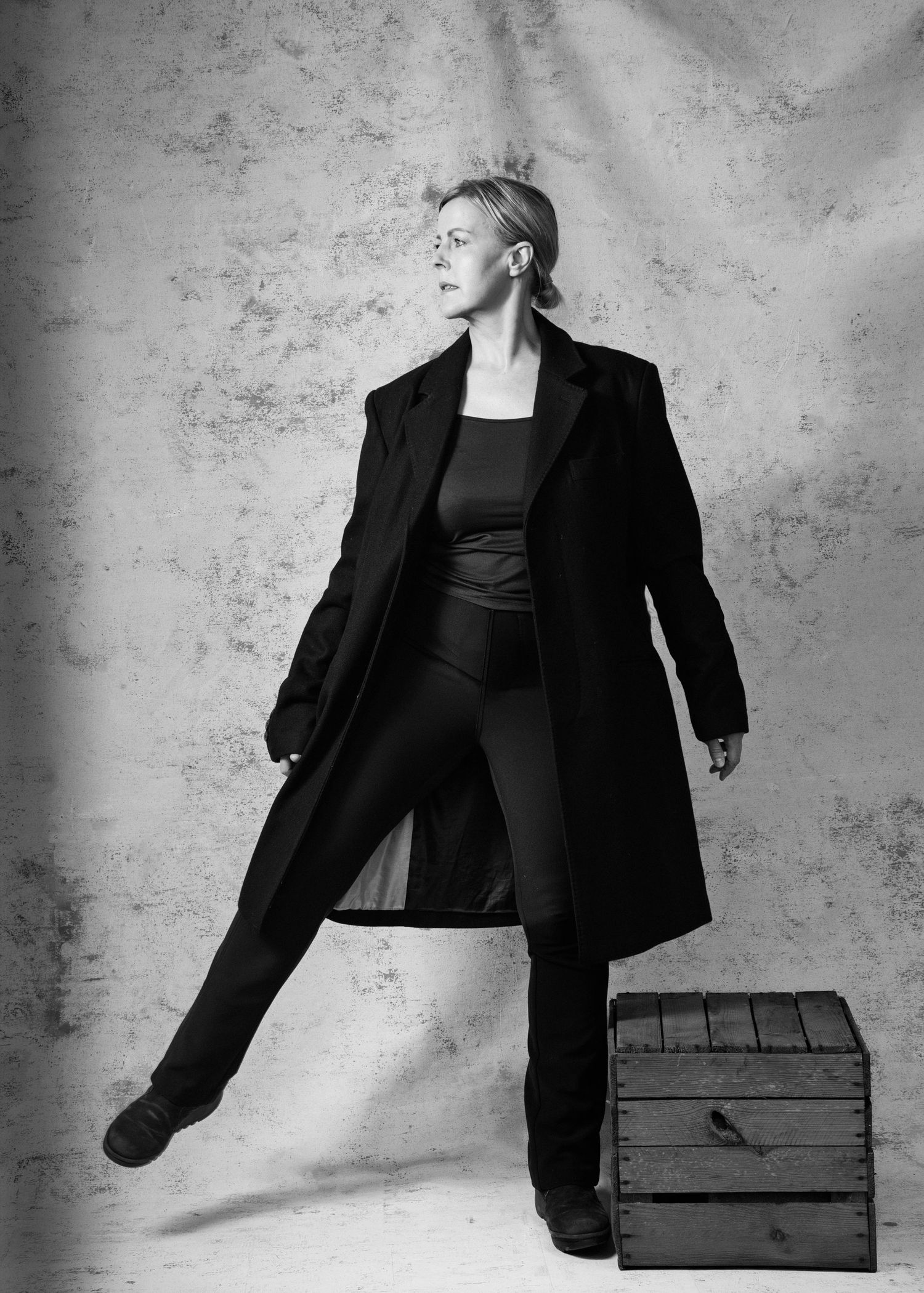 Black and white portrait of a woman in a long black coat posing confidently beside a wooden crate.