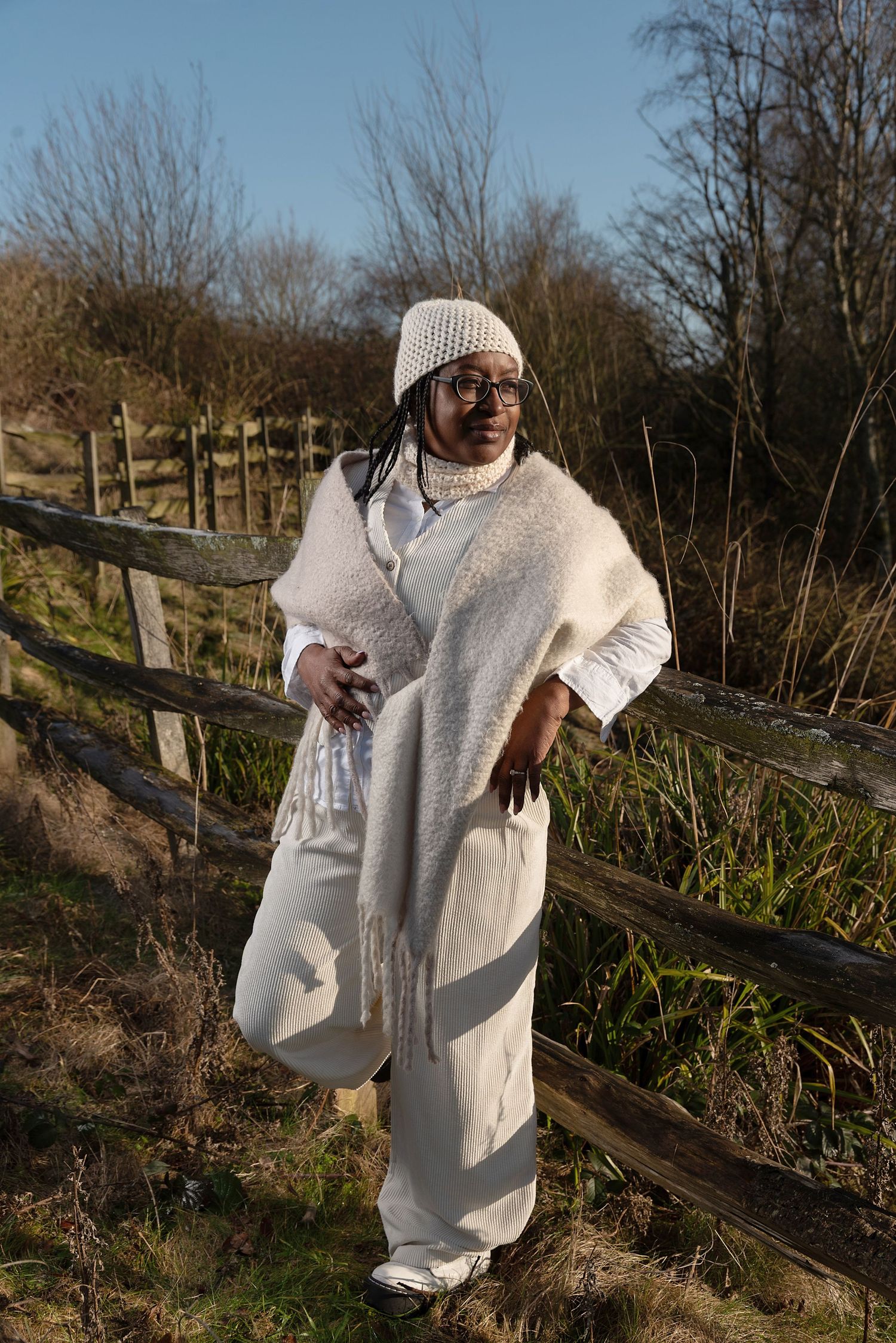 Someone wearing a cozy white winter outfit with knit beanie poses casually against a wooden fence in nature.