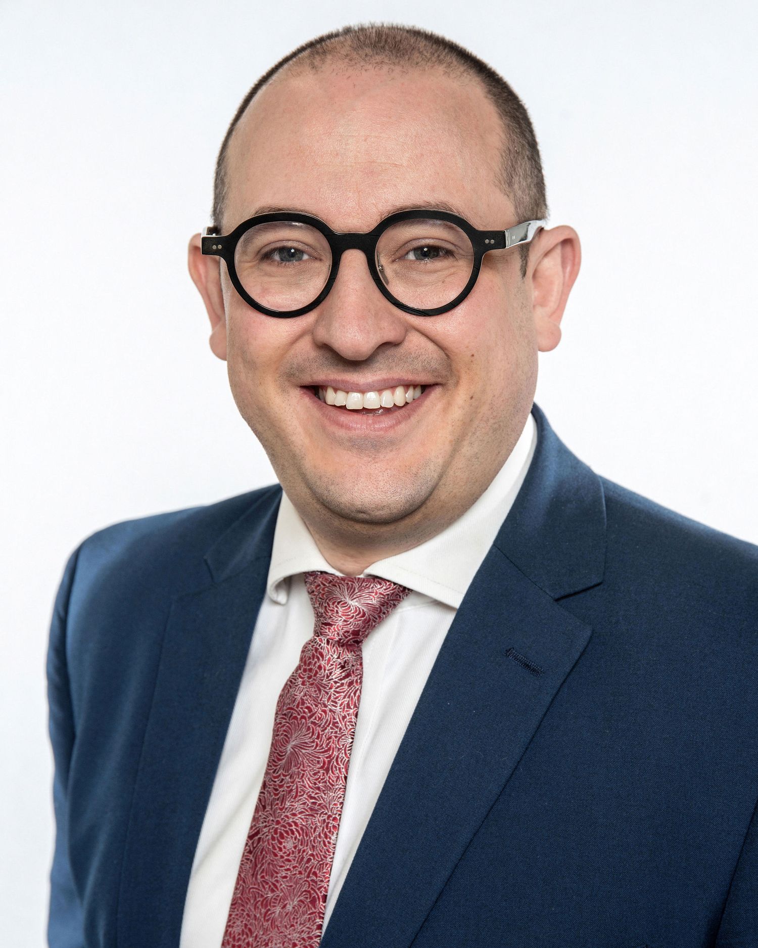 Professional headshot in navy suit with red tie and round glasses against white backdrop.