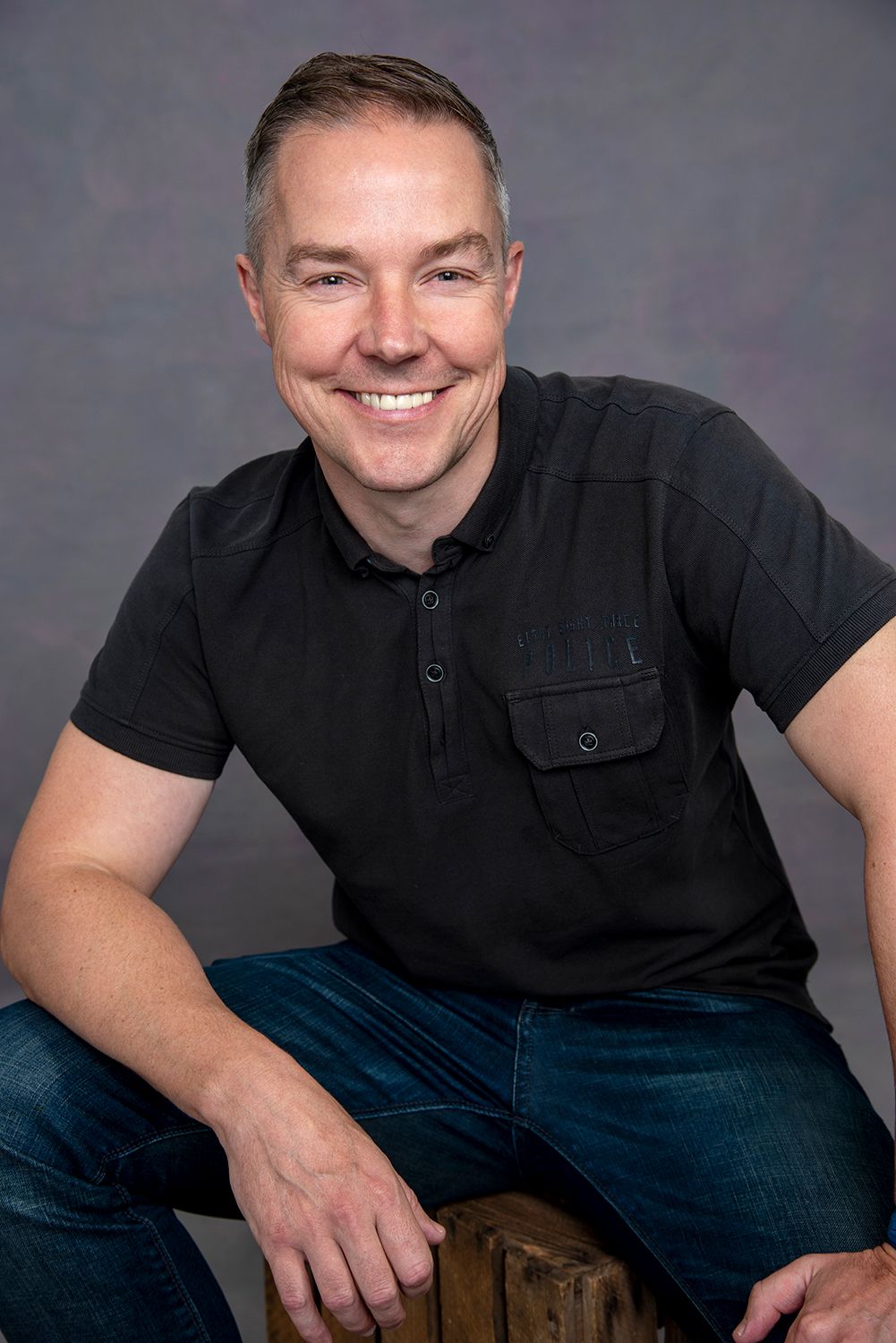 Professional studio portrait of person in black polo shirt and jeans seated casually against grey backdrop.
