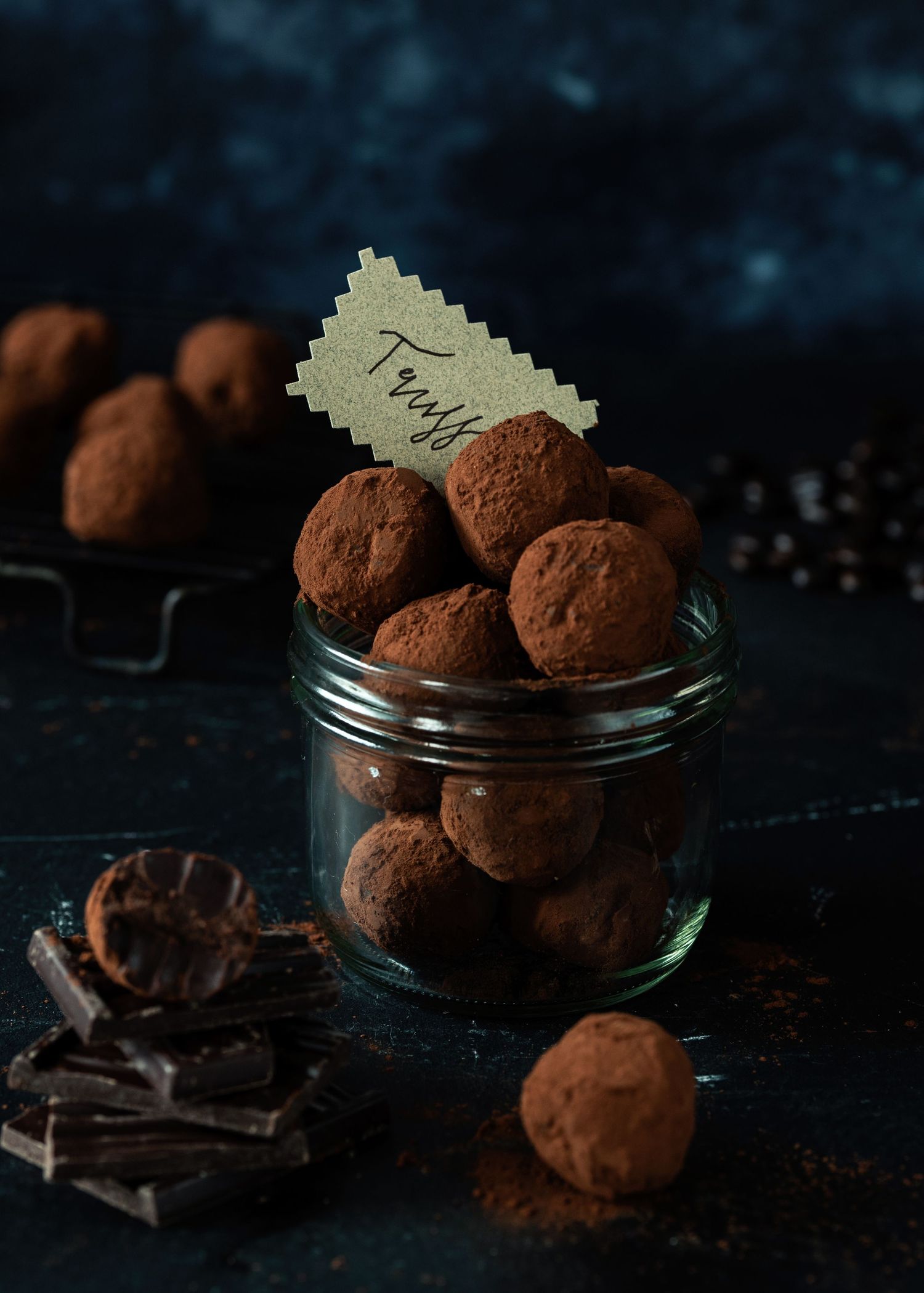 Glass jar filled with cocoa-dusted chocolate truffles labeled Trays, surrounded by dark chocolate pieces on a moody blue surface.