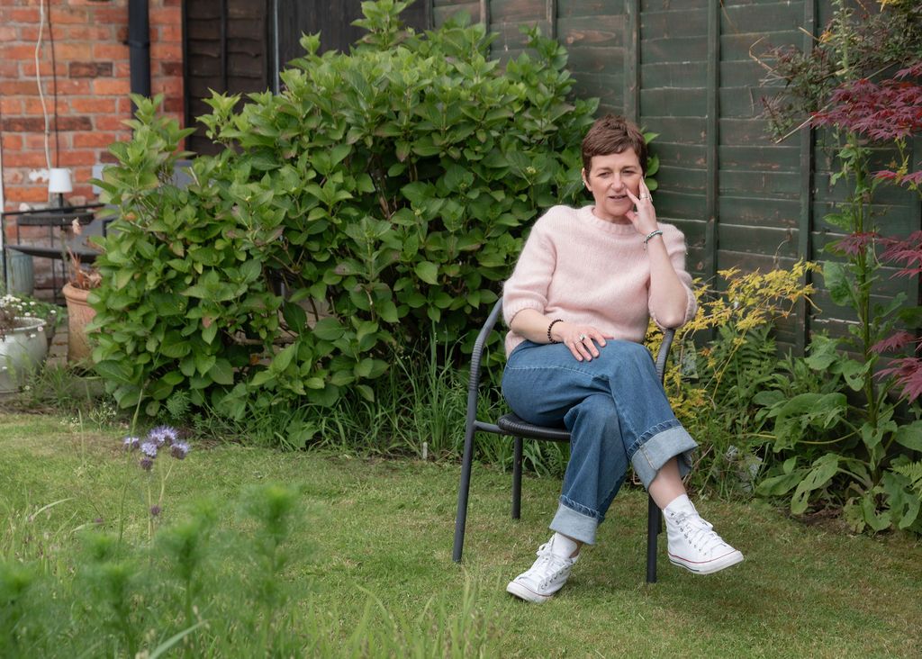 Woman in pink sweater and jeans sits on garden chair, relaxing outdoors surrounded by lush green shrubs and plants.