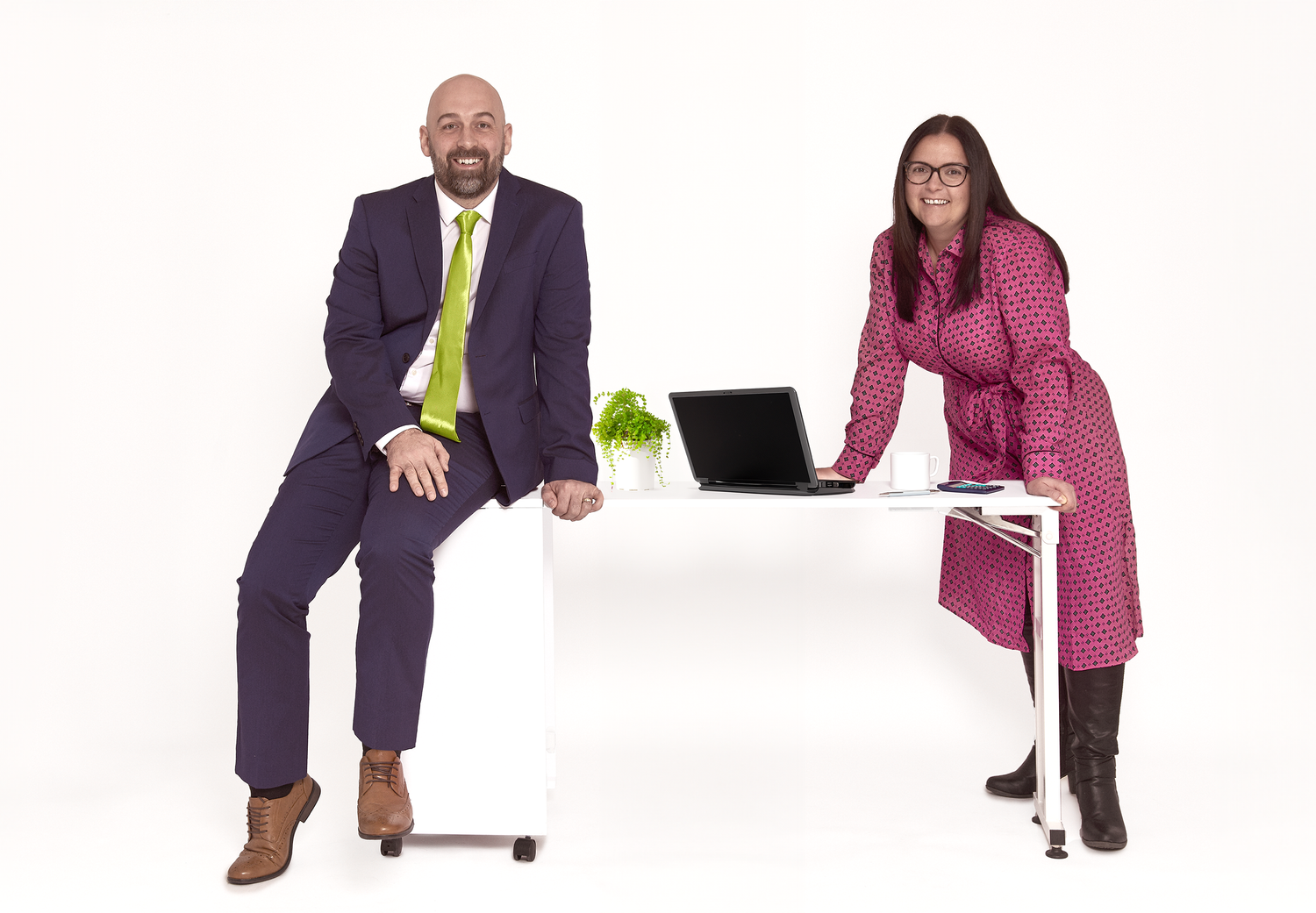 Two professionals pose at a modern white desk with a laptop in a business setting.