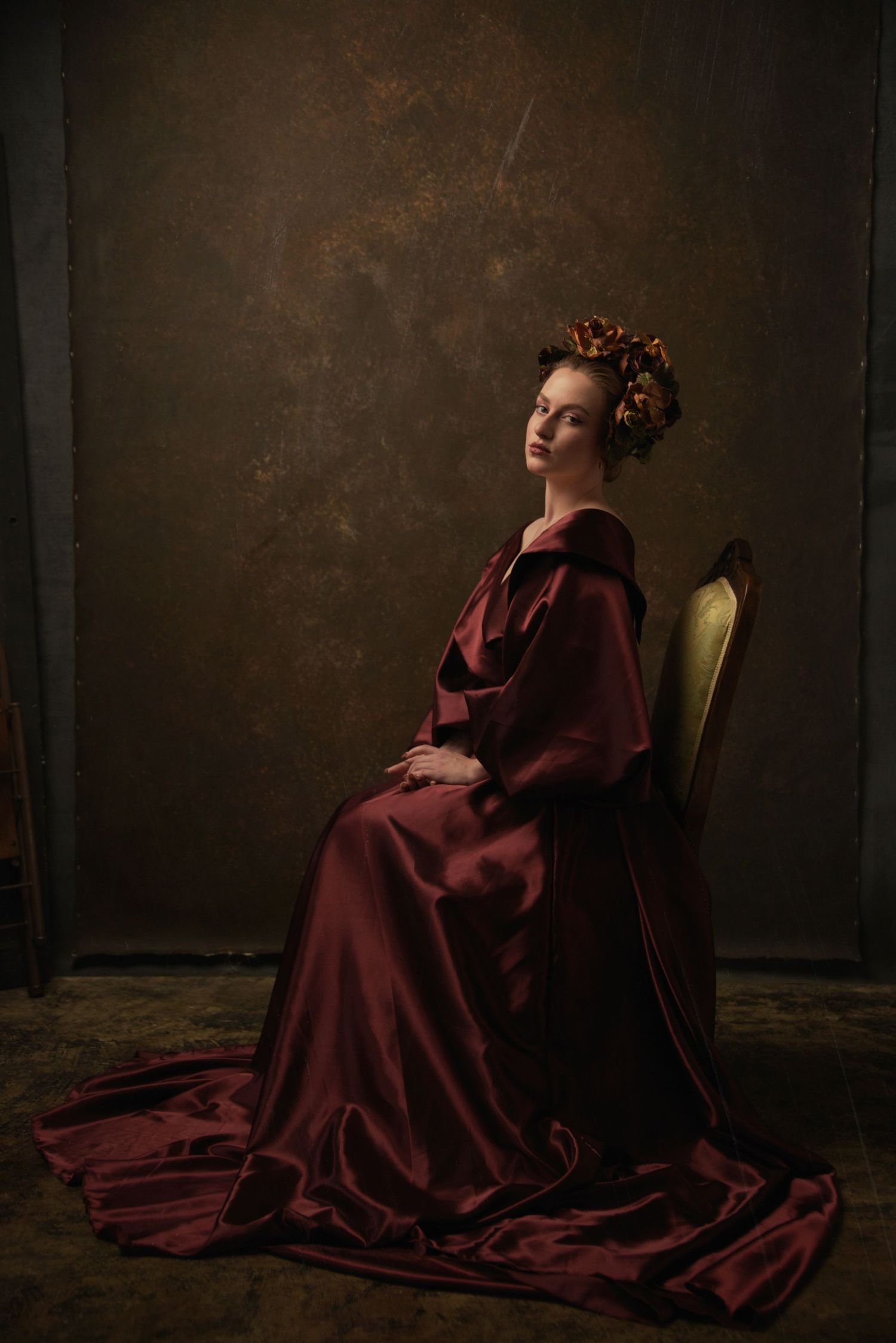 Elegant figure in flowing burgundy gown seated in vintage chair against dark backdrop.