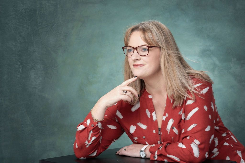 Person wearing glasses and red floral top poses pensively against textured green backdrop.