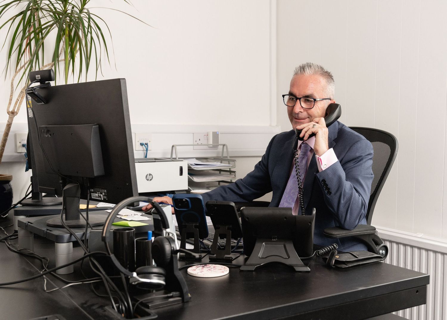 Person wearing glasses sits at desk with phone and computer in modern office environment.