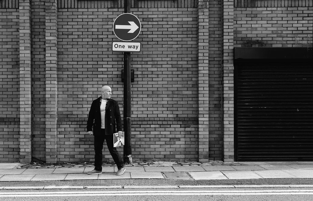 Black and white photo of a person standing under a one-way street sign in the opposite direction against a brick wall.