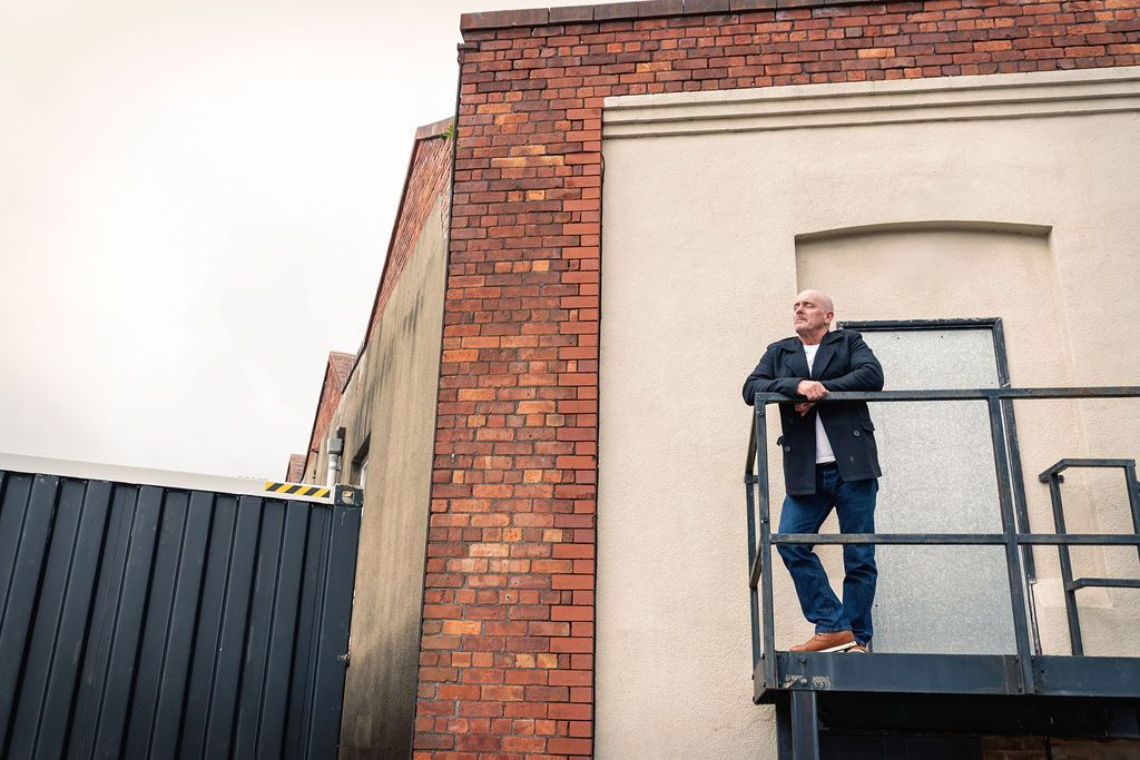 Man in black jacket stands on metal staircase beside brick building, looking into distance on overcast day.