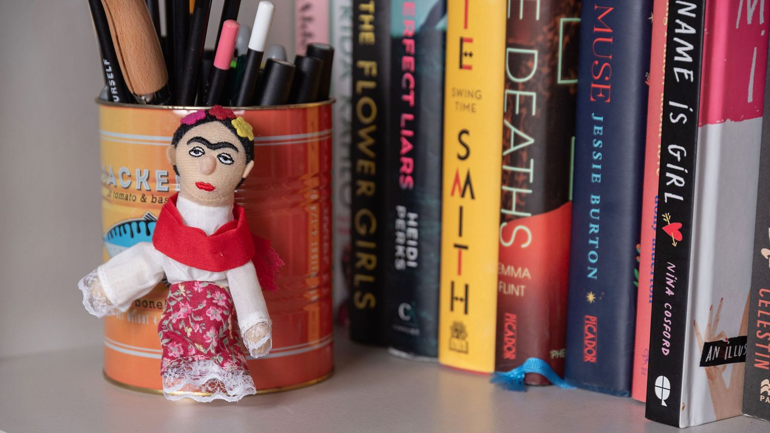Small plush doll in red dress sits in orange pencil holder next to row of colorful books on shelf.
