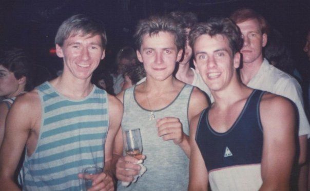 Three young men smiling together in a 1983 nightclub, wearing casual tank tops and holding a glass.