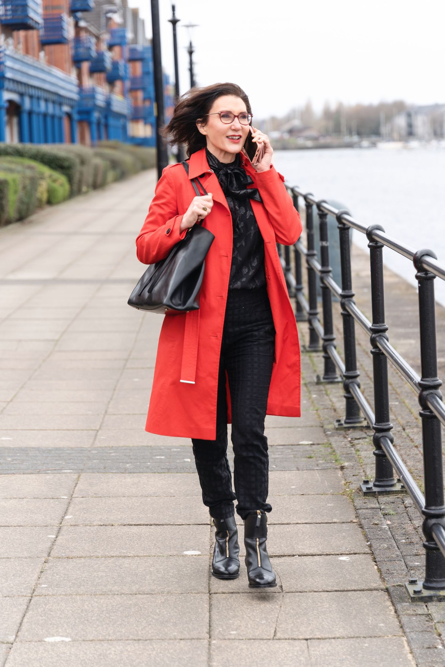 Woman in red coat and black outfit walks along waterfront path while talking on phone, carrying leather bag.