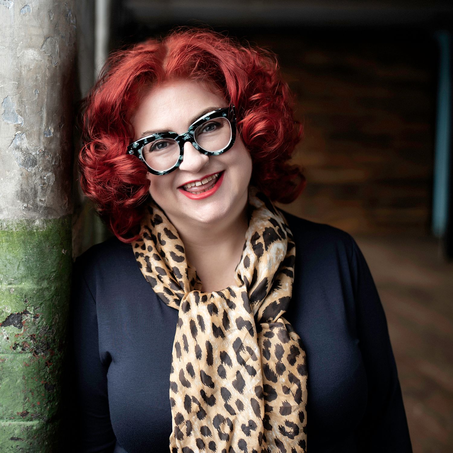Woman with red curly hair, leopard print scarf, and bold glasses smiling against a rustic background.