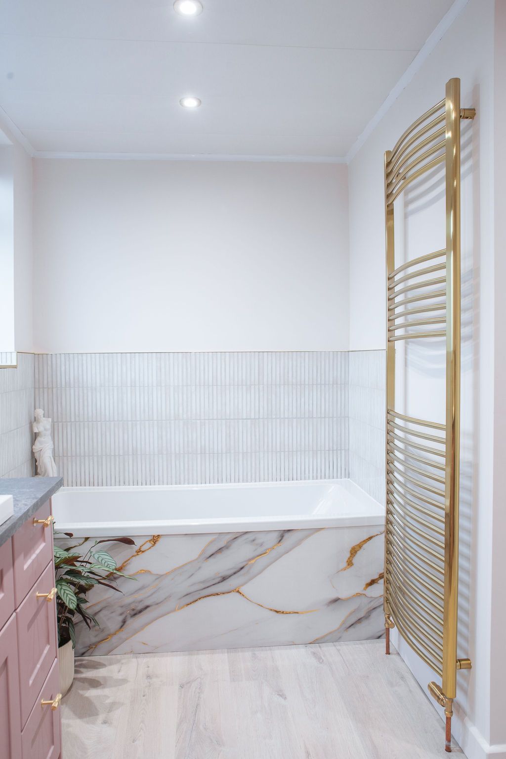 Modern bathroom with marble bathtub, gold towel warmer, and pink vanity features elegant white textured wall panels.