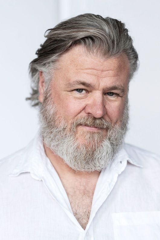 Portrait of a smiling man with gray hair and full beard wearing a white button-down shirt against light background.
