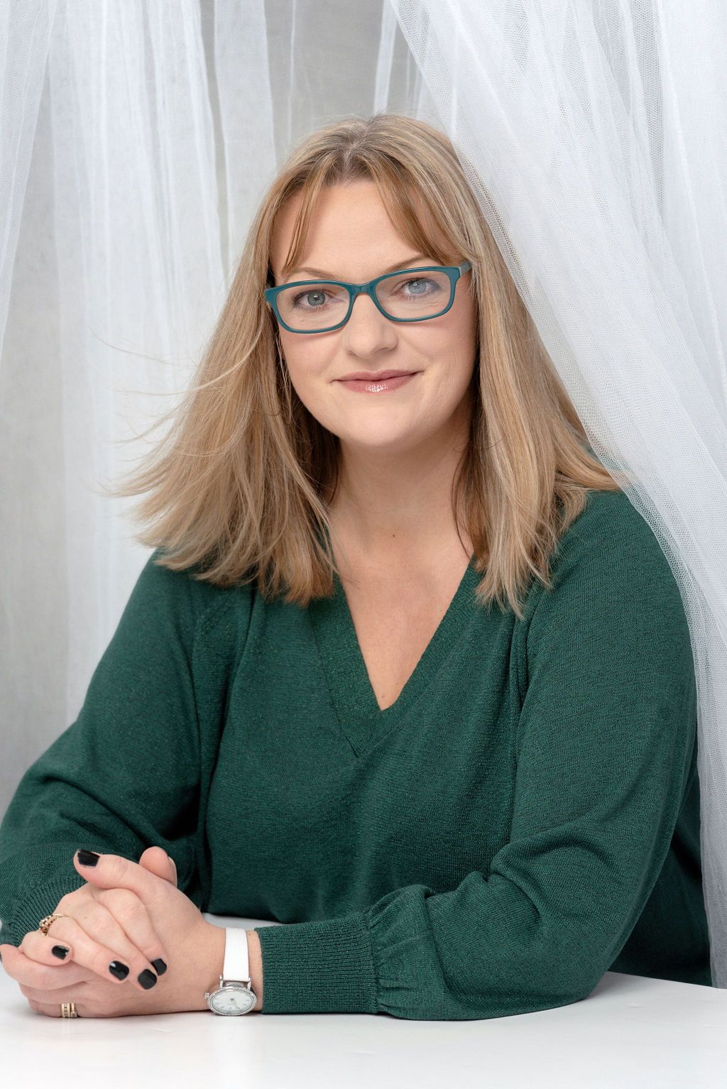 Posed portrait in green sweater and blue glasses against white curtained backdrop.