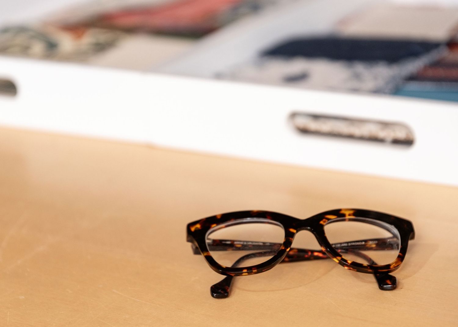 A pair of tortoiseshell eyeglasses resting on a beige surface with blurred background objects.