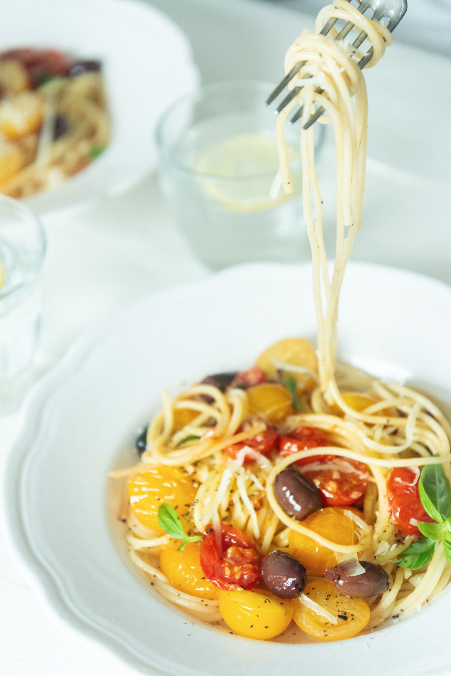 Fork twirling spaghetti with colorful cherry tomatoes and olives in a white bowl.