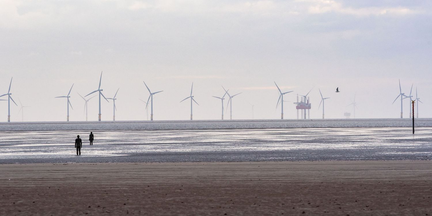 Two people walking on a sandy beach with offshore wind turbines and an oil platform visible on the horizon.