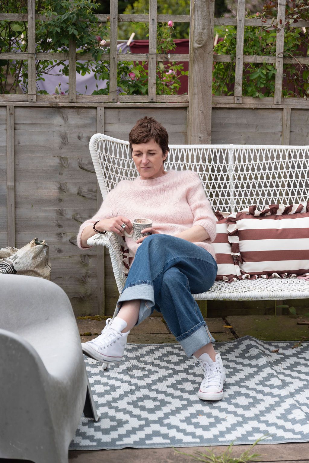 Woman relaxing on white garden bench with coffee, wearing pink sweater and jeans, on patio with geometric rug.