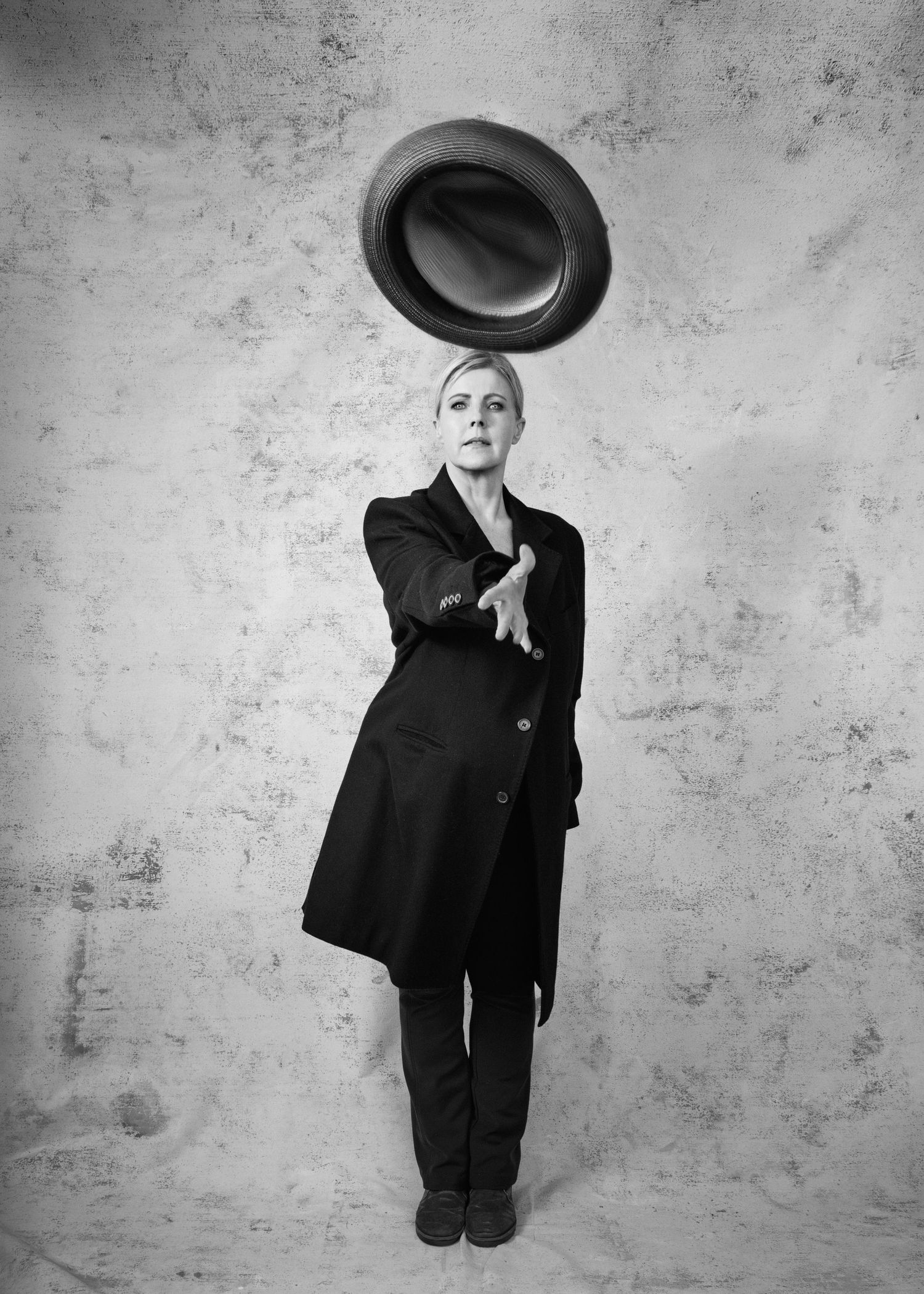 Black and white photo of a woman in a dark coat tossing a hat into the air against a textured background.
