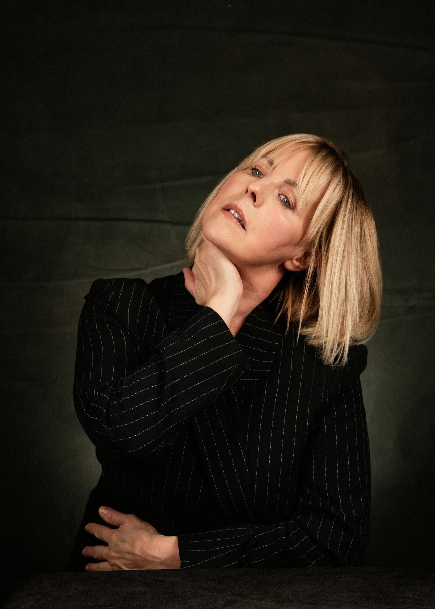 Blonde woman in black pinstripe blazer poses dramatically, hand on neck, looking upward against dark background.