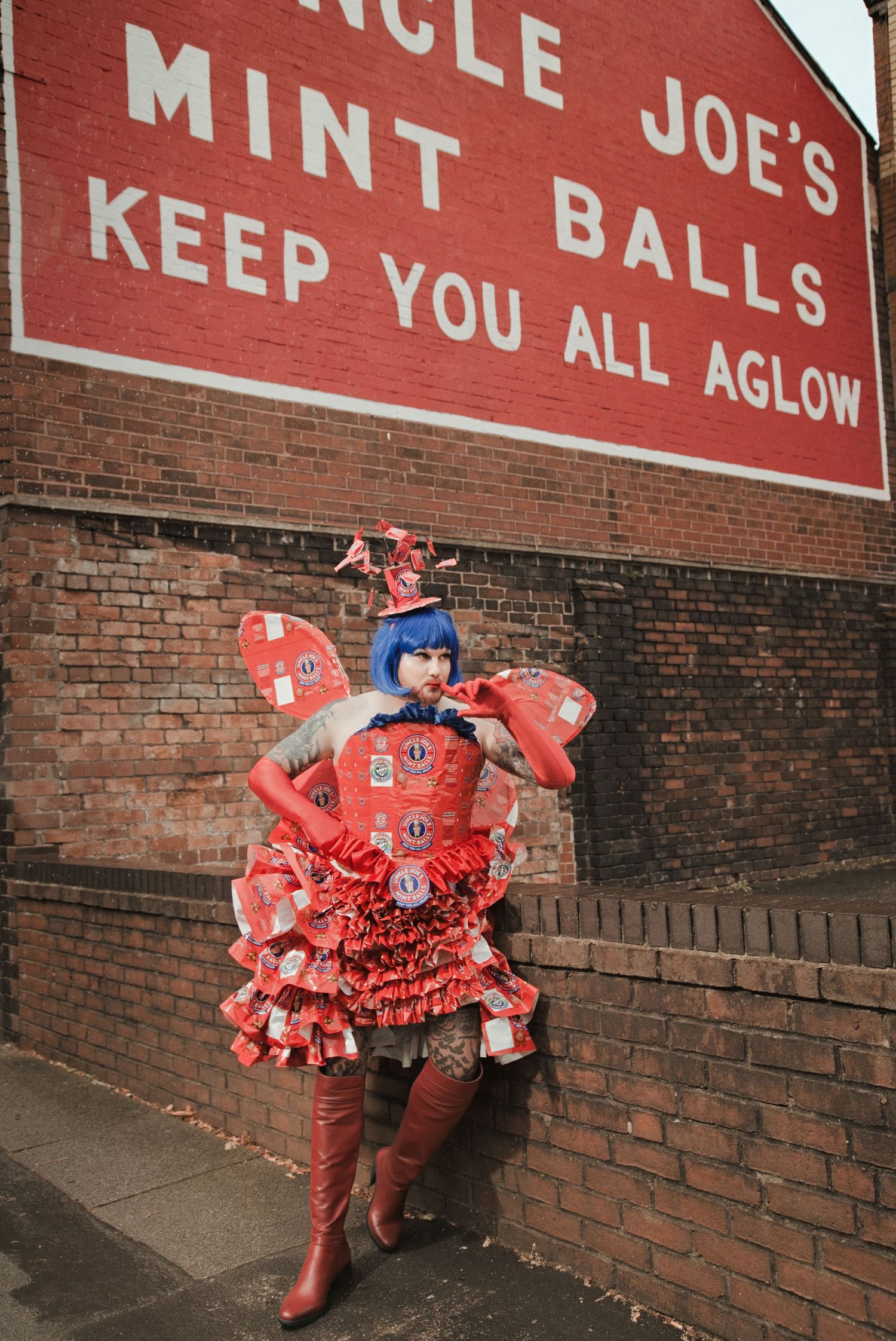 Person in red costume and blue wig poses by Uncle Joe's Mint Balls brick wall advertisement.