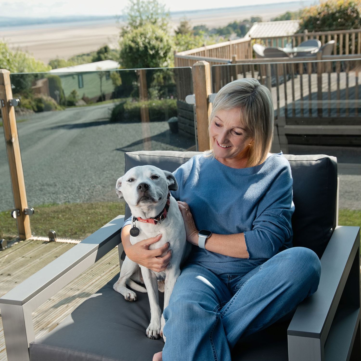 A smiling blonde woman sits outdoors on a patio chair, lovingly holding a white dog with a red collar.