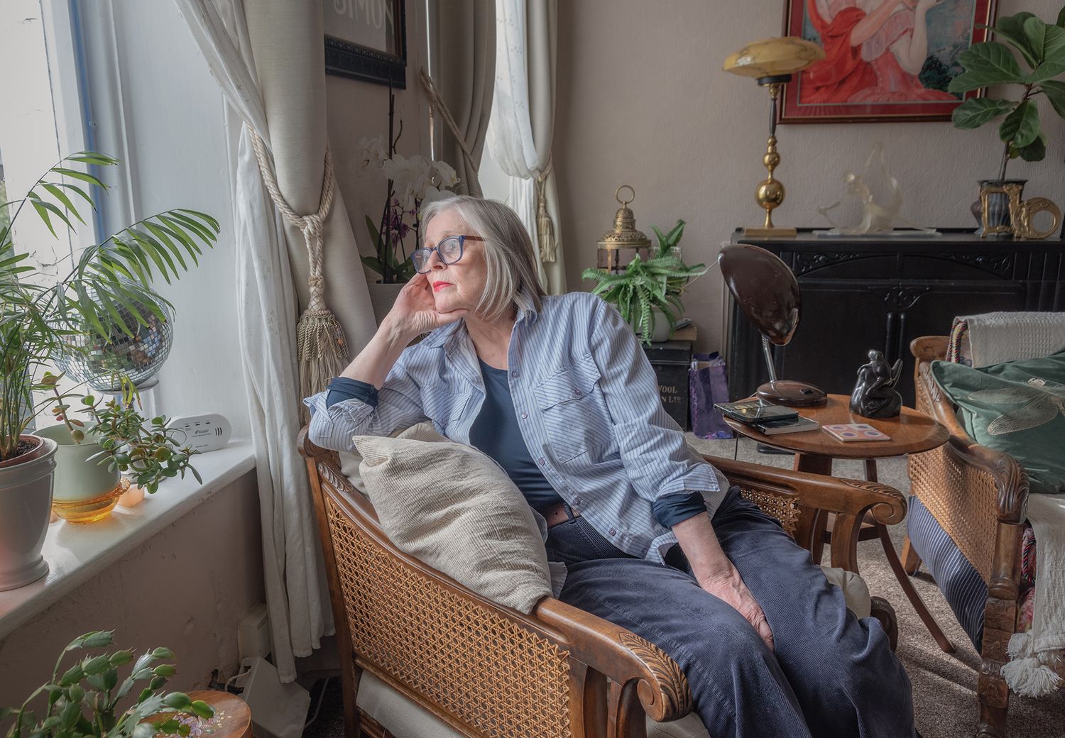 Elderly woman with gray hair sits thoughtfully in a wicker chair by a window, surrounded by plants and eclectic home decor.