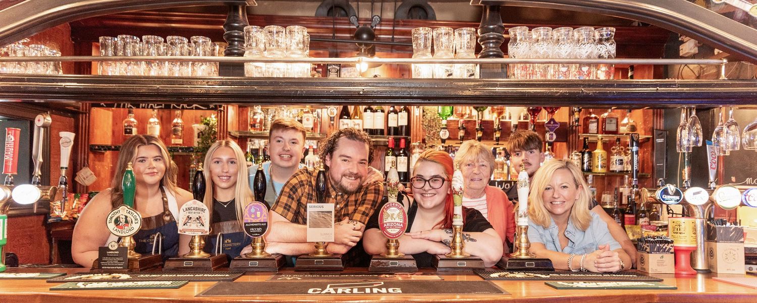 Group of cheerful bar staff posing behind a pub counter with beer taps and fully stocked shelves in background.