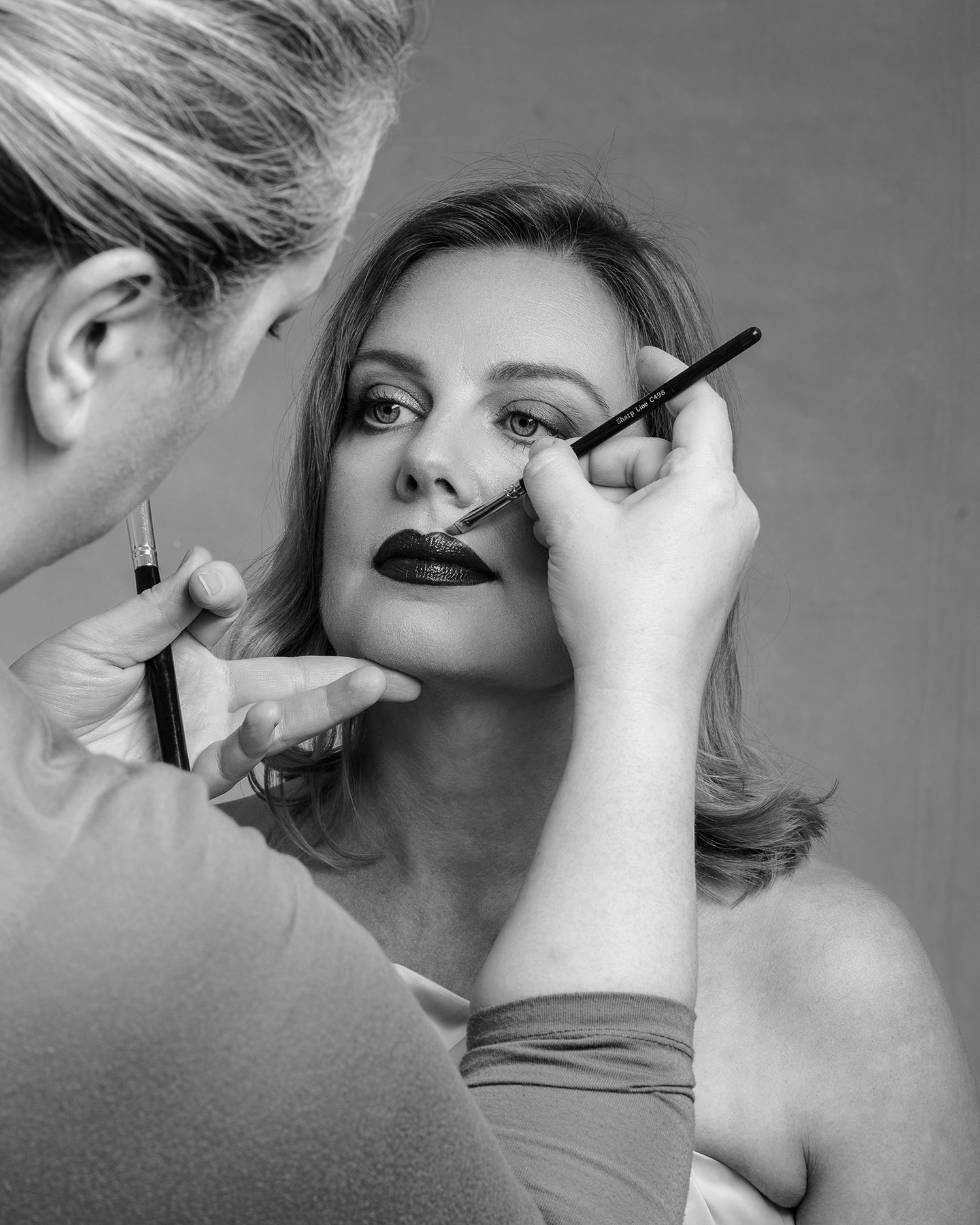 Black and white photo of a makeup artist applying eyeliner to a woman with dark lips during a professional session.