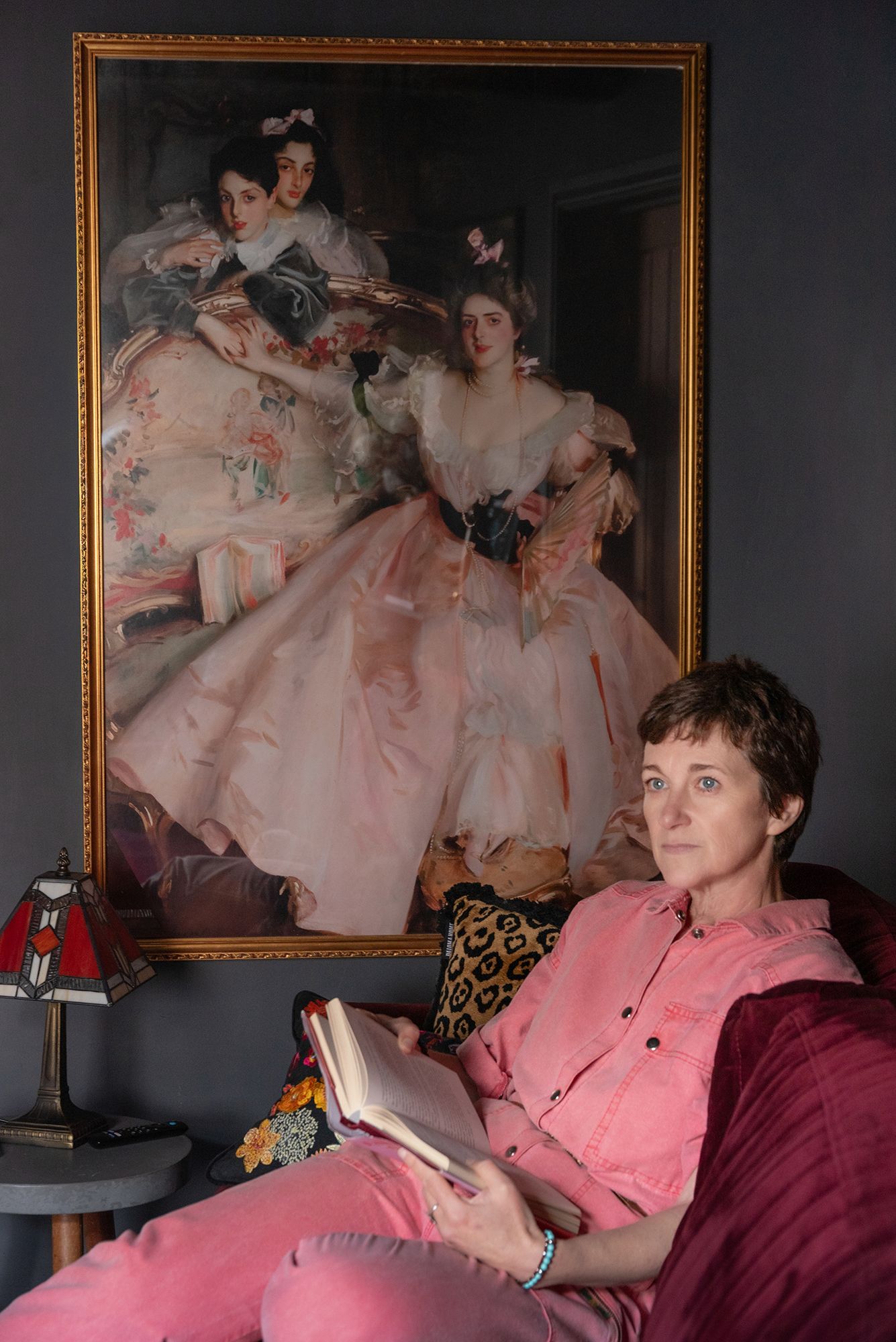 Woman in pink reading a book on a sofa beneath a framed classical portrait painting in an elegant room.
