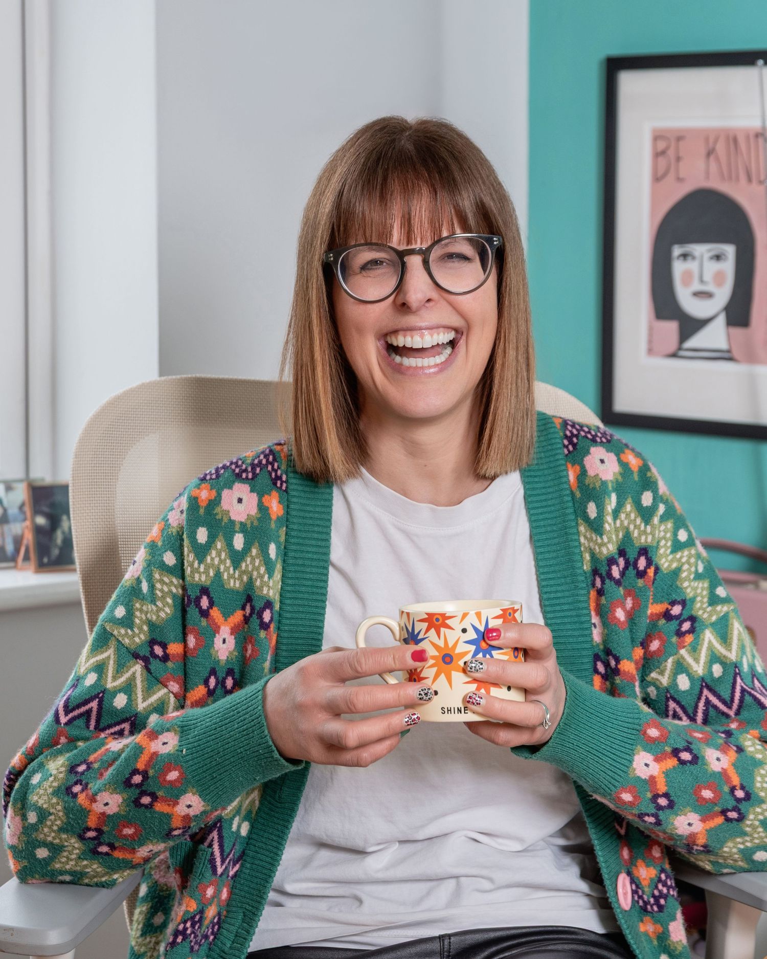 A smiling woman with glasses and a colorful floral cardigan holds a decorative mug while sitting in a bright, cheerful room.