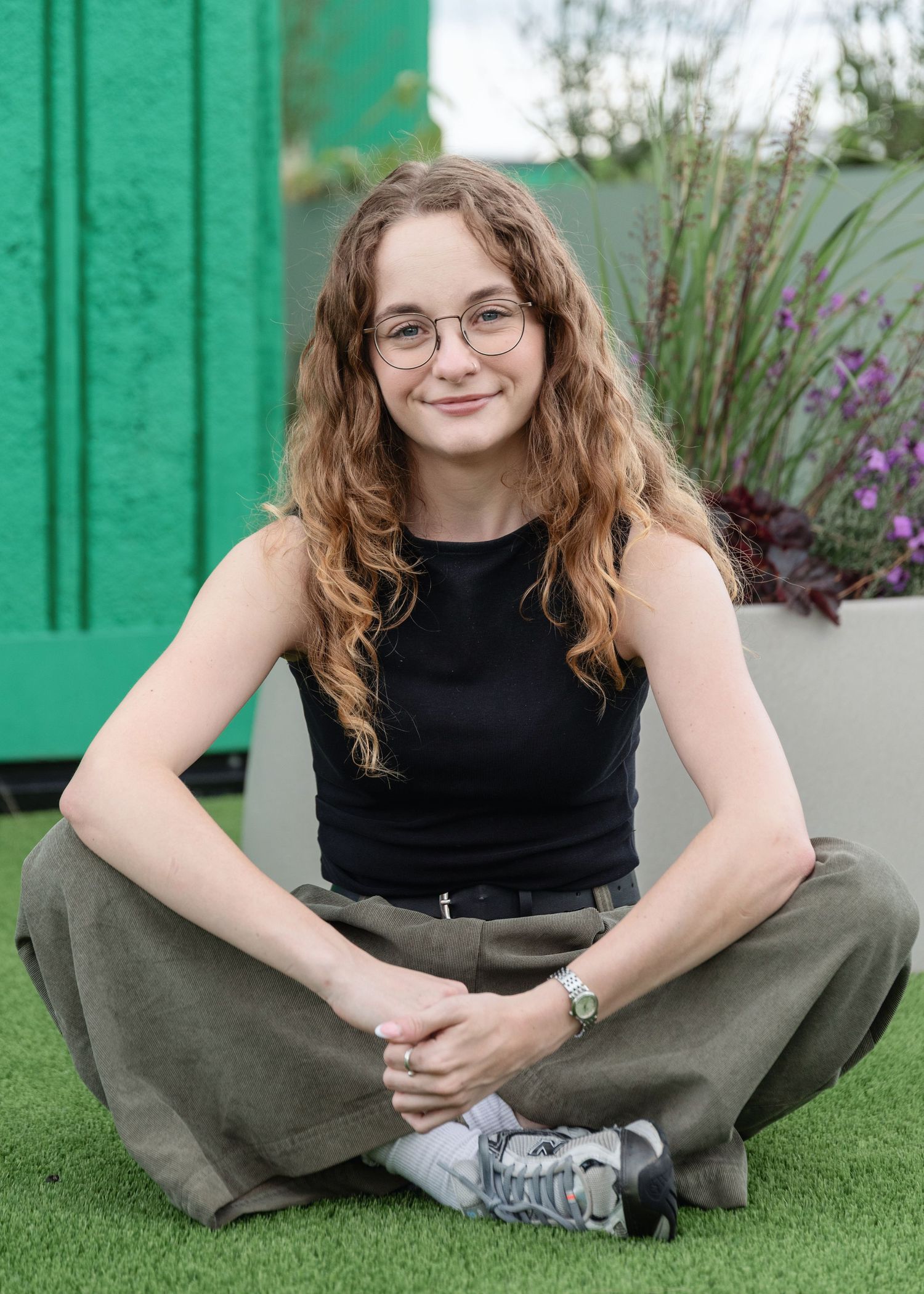 Young woman with curly hair and glasses sitting cross-legged on grass in a black top and olive pants.