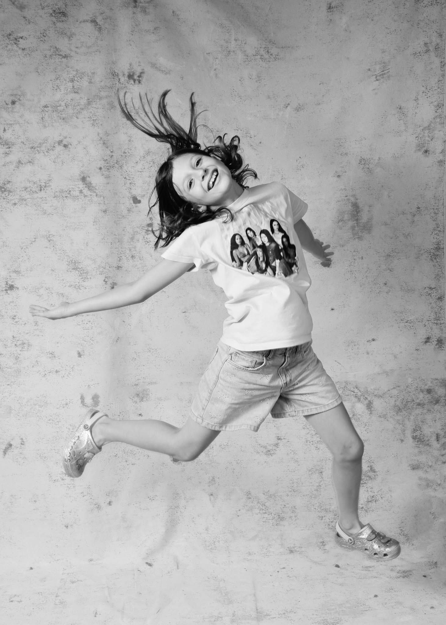 Black and white photo of a joyful young girl jumping mid-air with hair flying, wearing a white t-shirt and denim shorts.