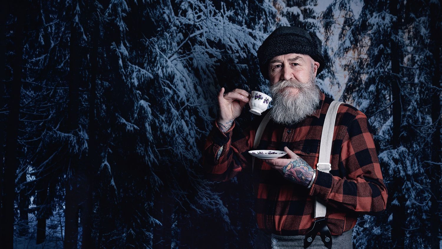 A person in a plaid shirt and black hat drinks from a mug while sitting outdoors in a snowy winter forest.