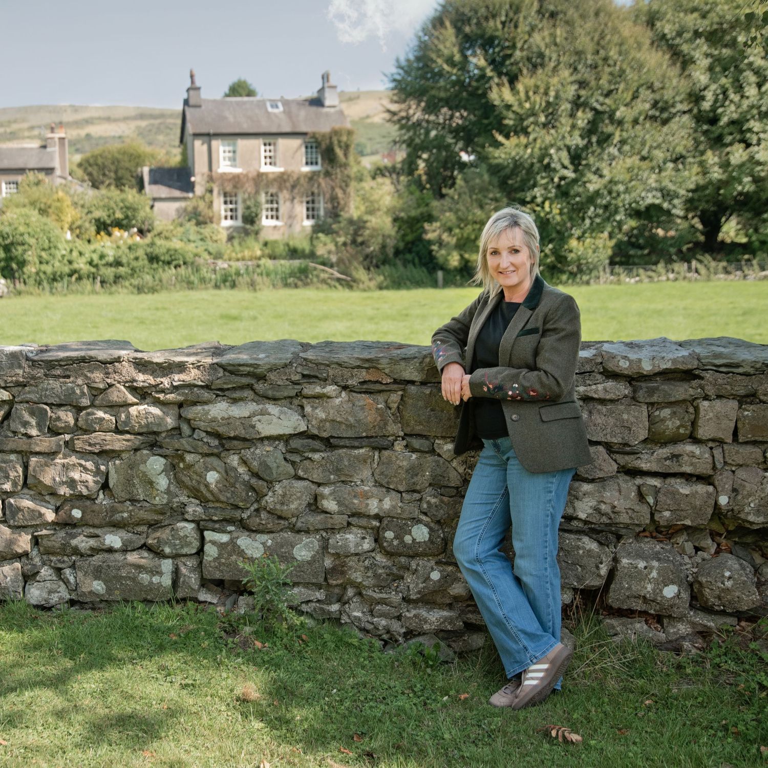 Woman in blazer leaning against a rustic stone wall with a countryside house and green lawn behind her.