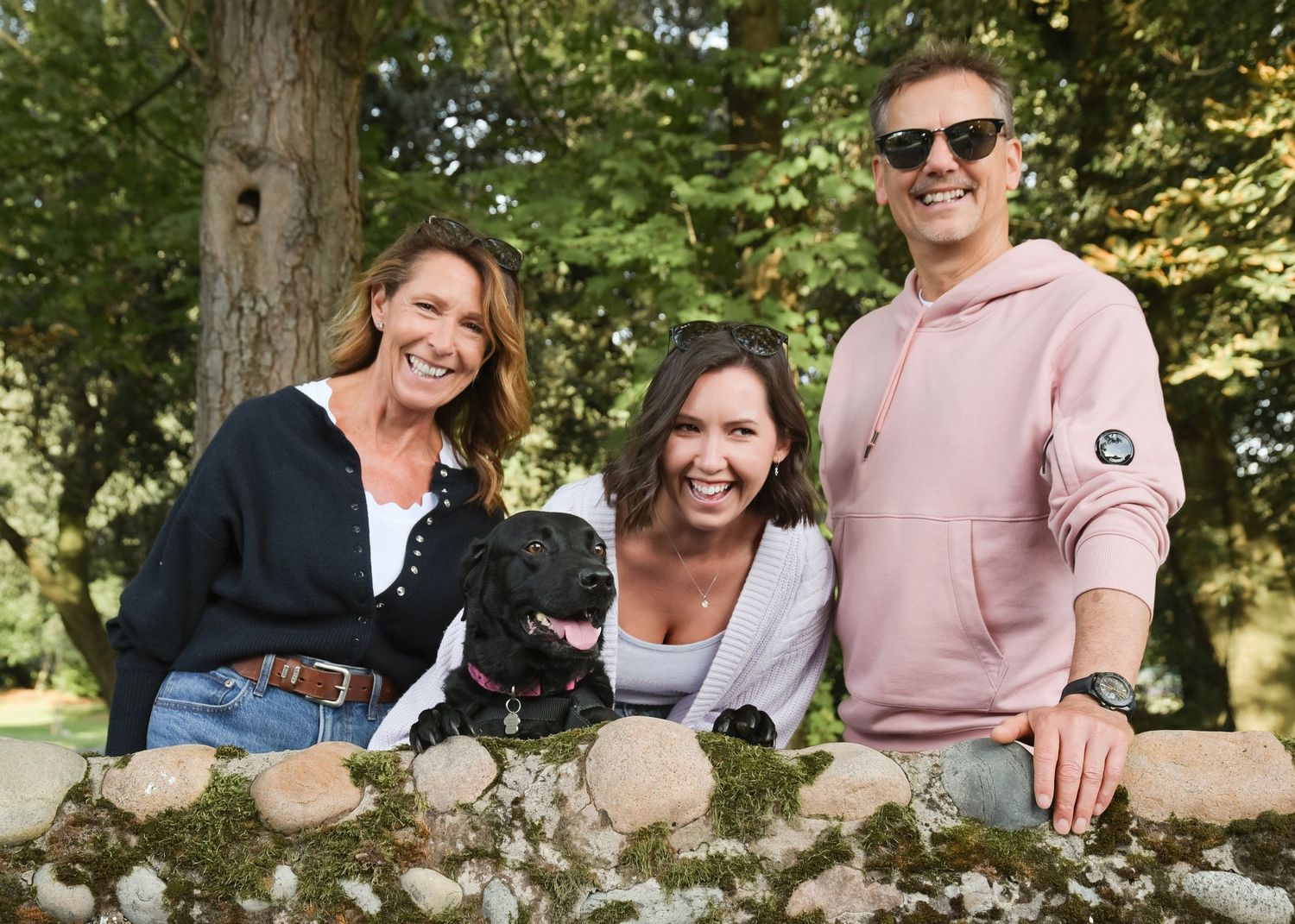 Two women and a man smile outdoors with a black dog, posing by a mossy stone wall in a lush green park.