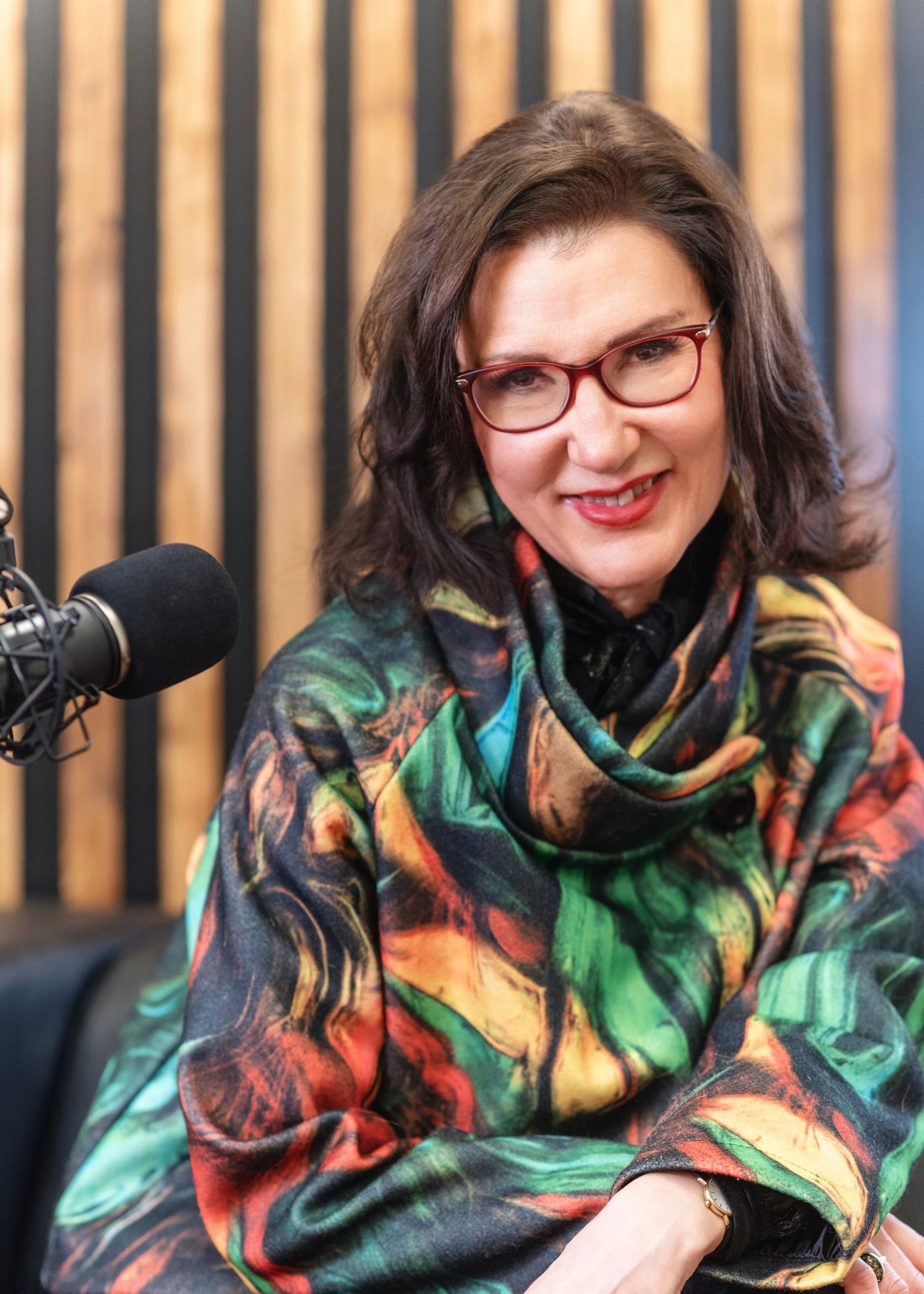 Professional wearing colorful patterned scarf and glasses speaks into podcast microphone against wooden paneled backdrop.