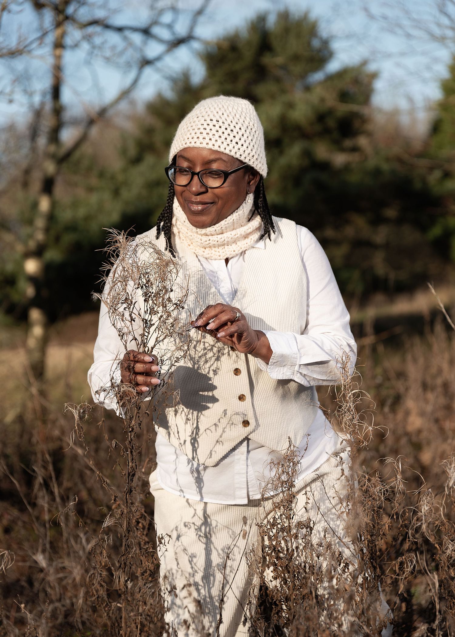 A person wearing a white knit hat and glasses examines dried plants in a natural outdoor setting during winter.