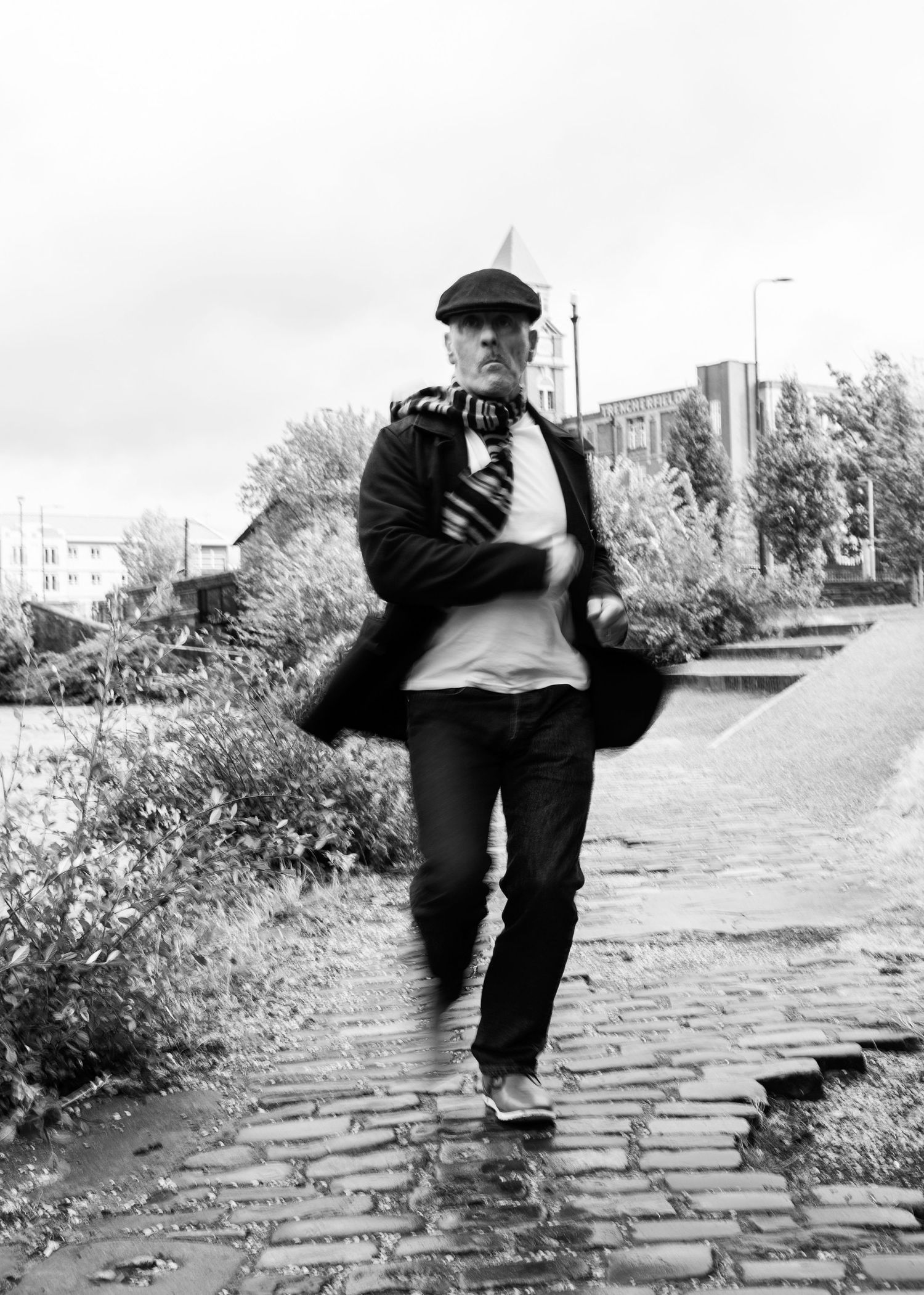 Black and white photo of someone in a flat cap and coat jumping on a cobblestone path on an overcast day.