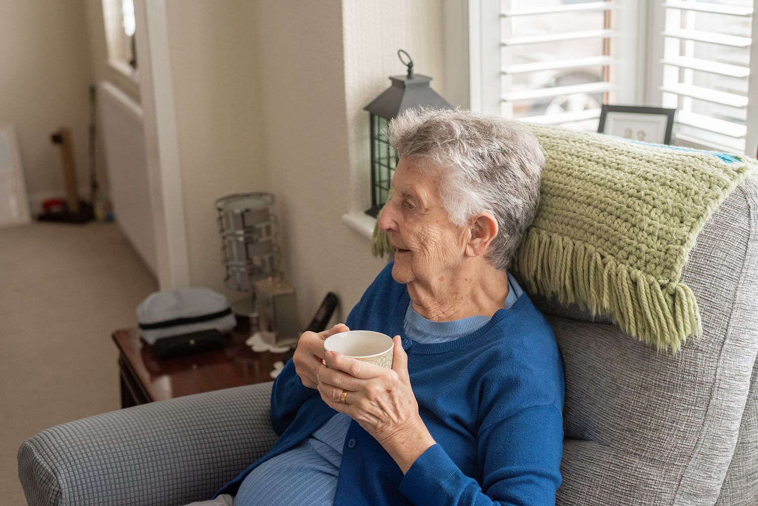 Elderly woman sitting in armchair holding a mug, gazing thoughtfully in a cozy living room.