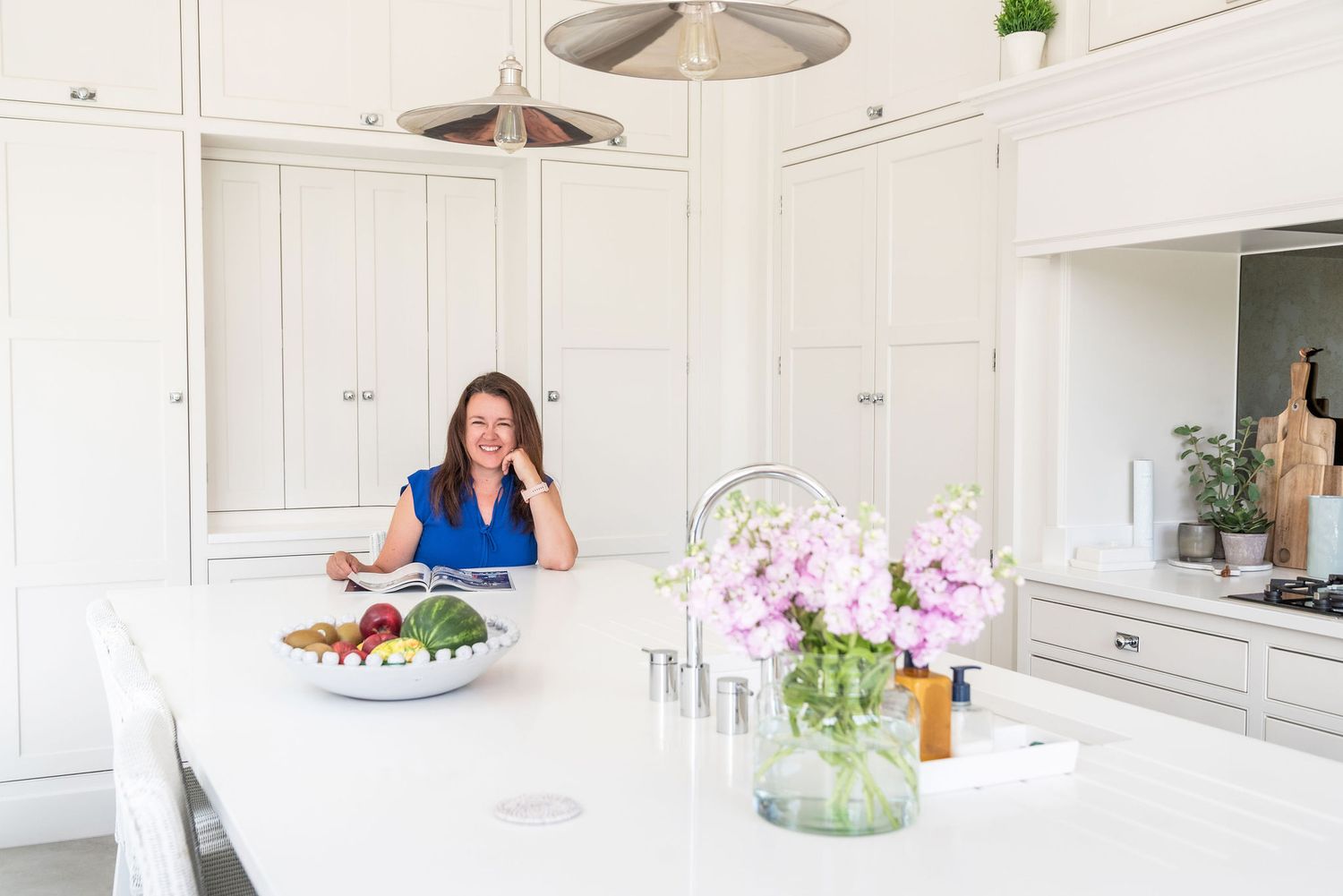 Woman smiling in a bright white kitchen with fresh vegetables and pink flowers on a marble island.