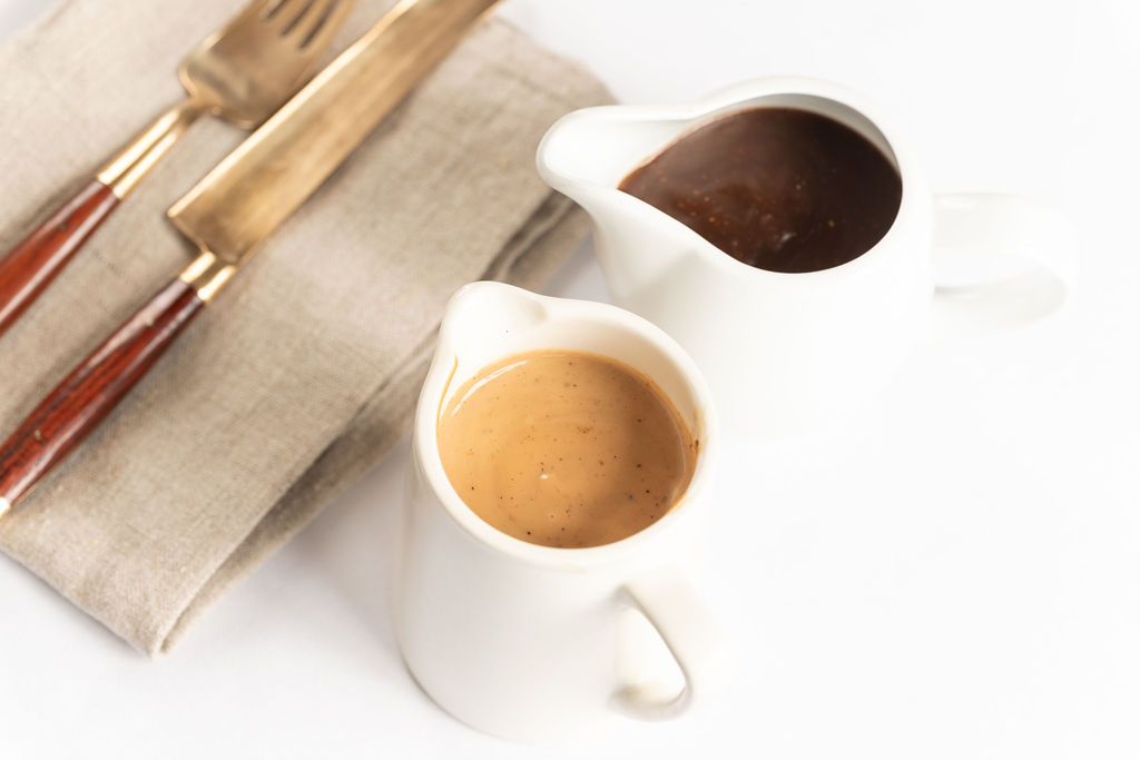 Two white ceramic pitchers with coffee and chocolate sauce beside vintage cutlery on a linen napkin.