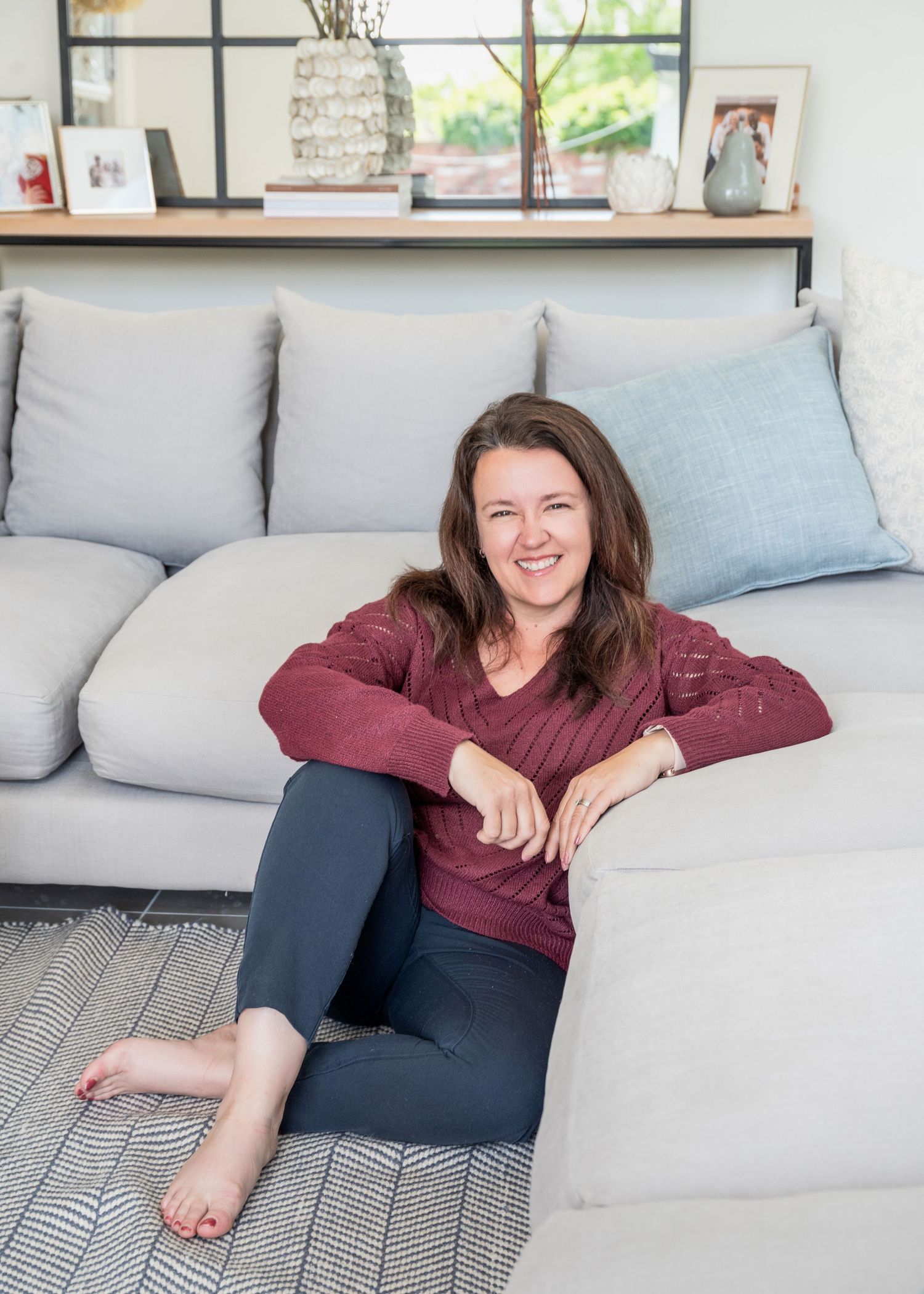 Smiling woman in burgundy top and gray pants sitting barefoot on floor leaning against light gray sofa.