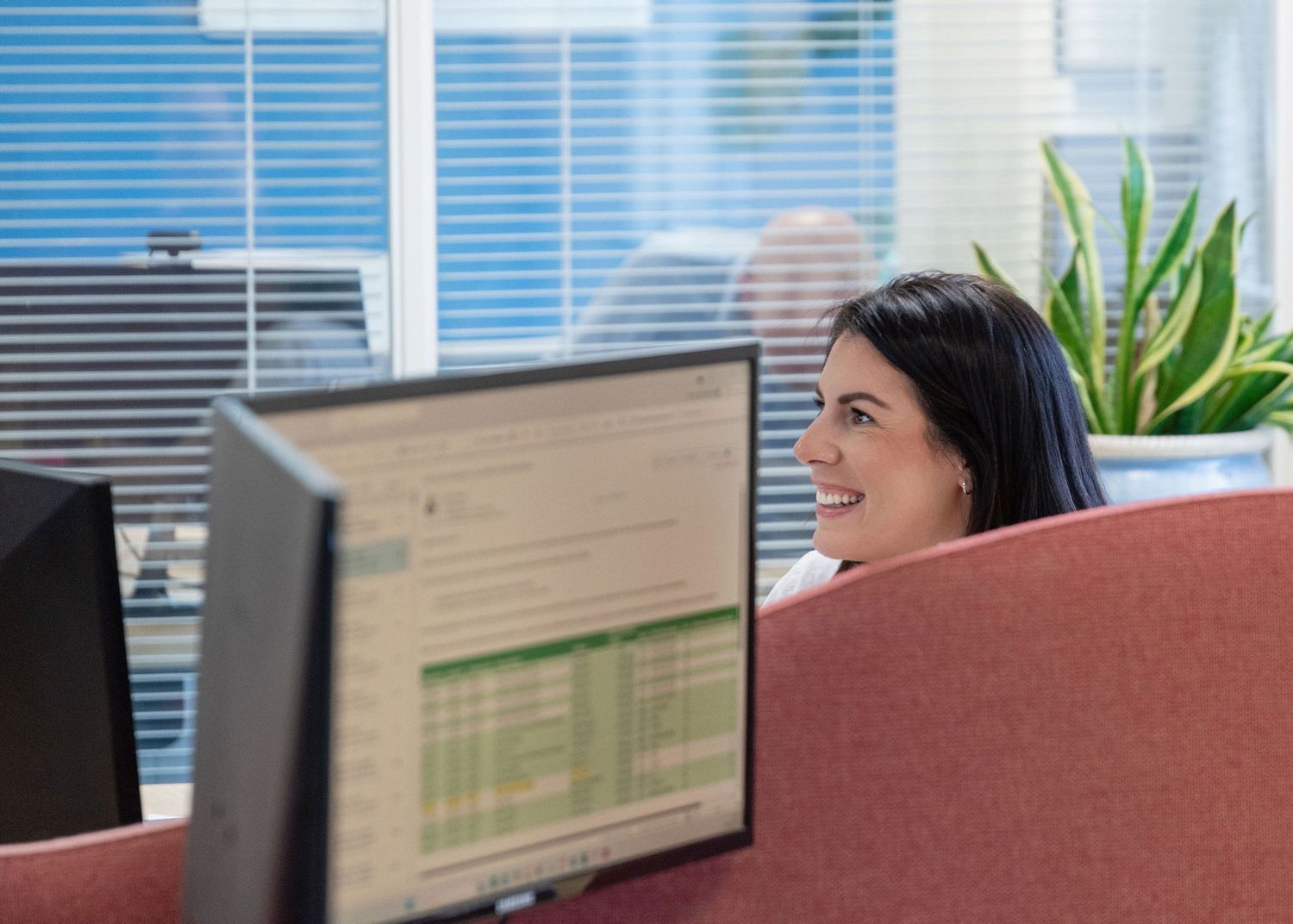 Smiling woman working at a computer workstation in a modern office cubicle with green plants and blinds.