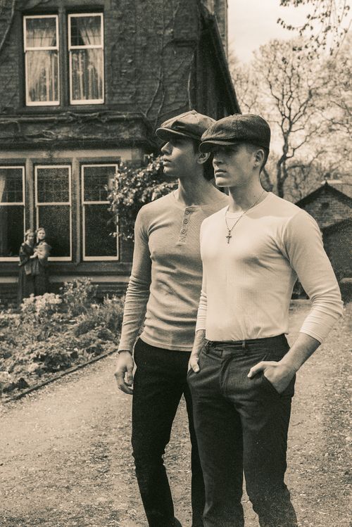 Two young men in flat caps and long-sleeve shirts pose outdoors near a vintage house in a sepia-toned photo.