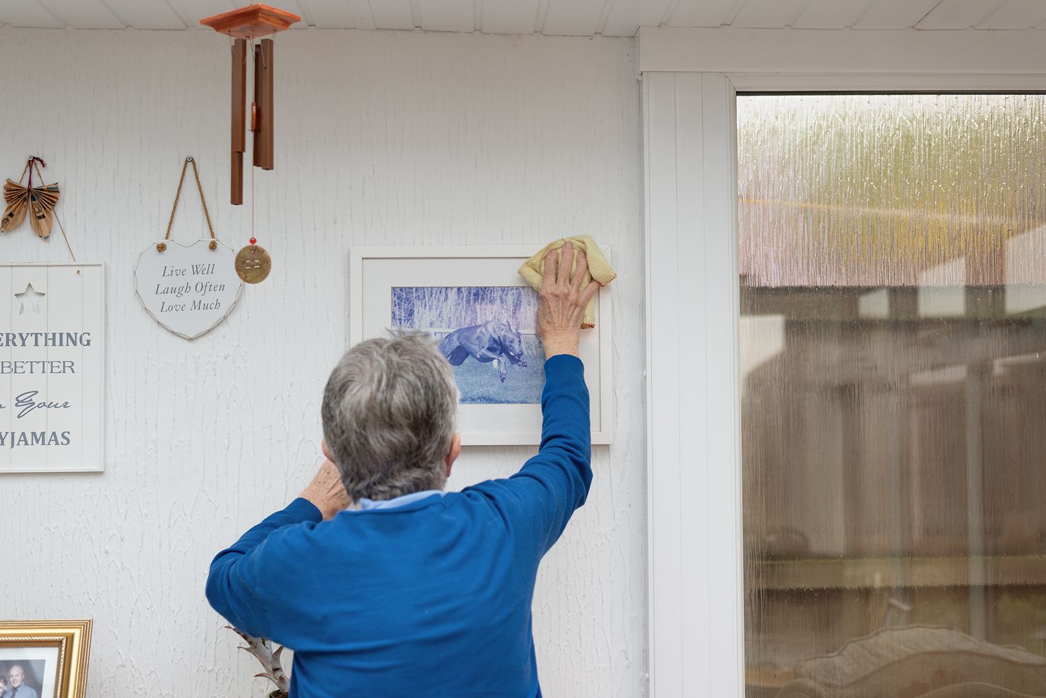 Elderly woman in blue sweater cleaning a framed picture on a white wall next to a window.