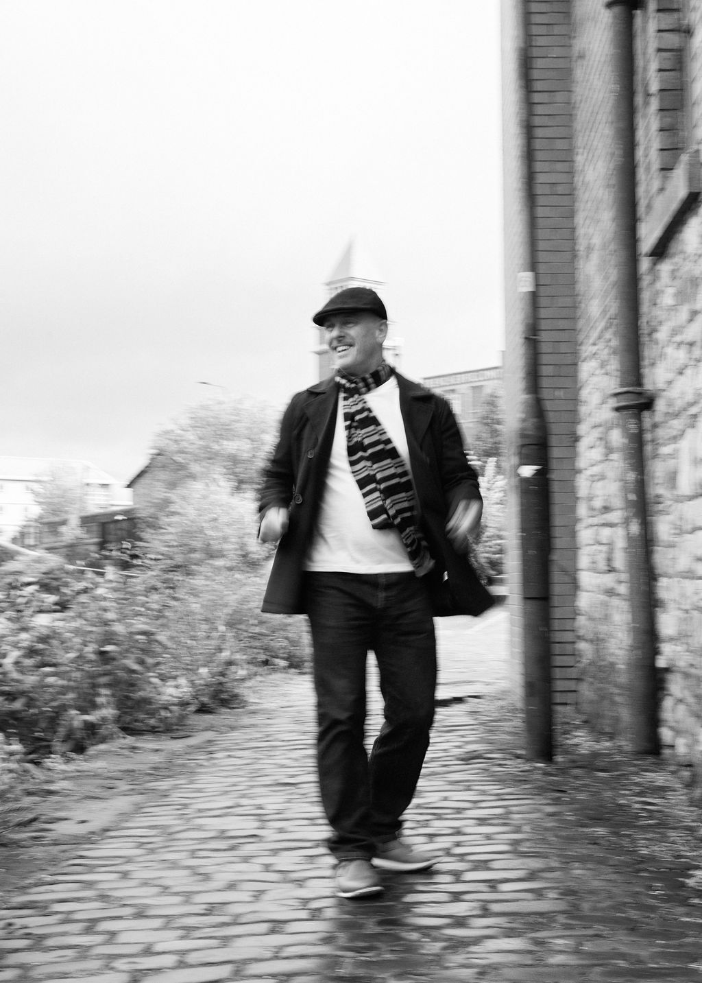 Man in flat cap and striped scarf walks confidently on cobblestone path beside brick building in black and white photo.
